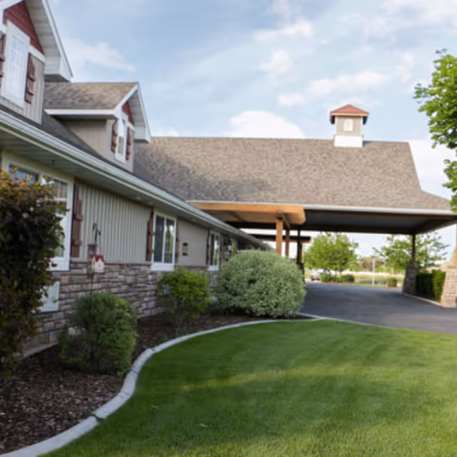 Exterior view of The Gables Memory Care of Shelley building with a covered entrance, manicured lawn, shrubs, and a partly cloudy sky.