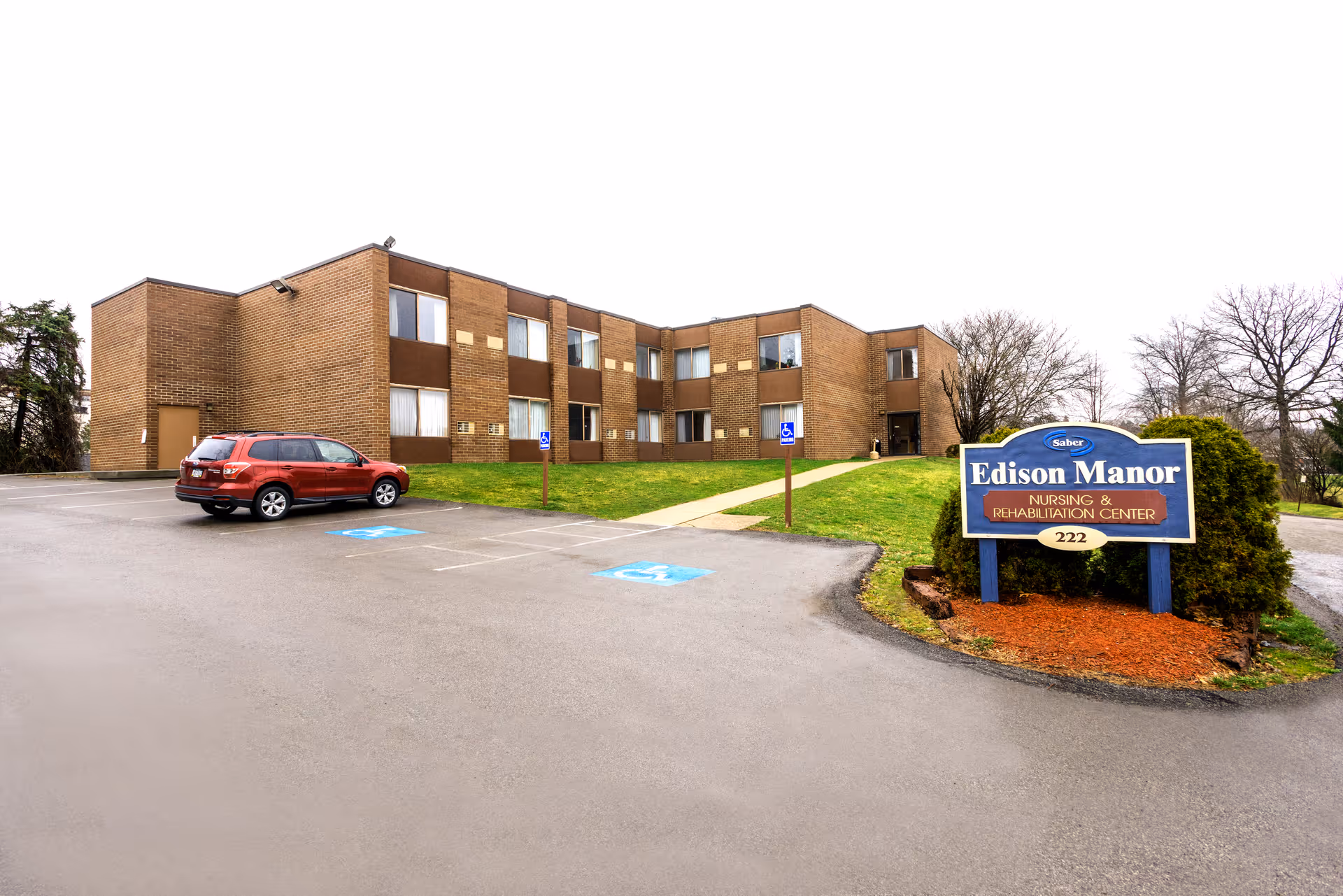 Exterior view of Edison Manor Nursing and Rehabilitation Center, a two-story brick building with several windows. There is a parking lot in front with a red SUV parked and two handicap parking spaces marked. A blue and white sign with the facility's name and address is visible on the right side near some bushes.