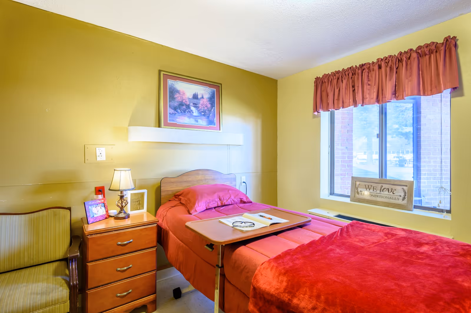 A cozy bedroom in a senior living facility with a single bed covered in red bedding and a wooden overbed table holding a book and glasses. Next to the bed is a wooden nightstand with a lamp, framed photos, and a small decorative sign. A cushioned chair is positioned beside the nightstand. The room has yellow walls, a window with red curtains, and a framed landscape painting above the bed.