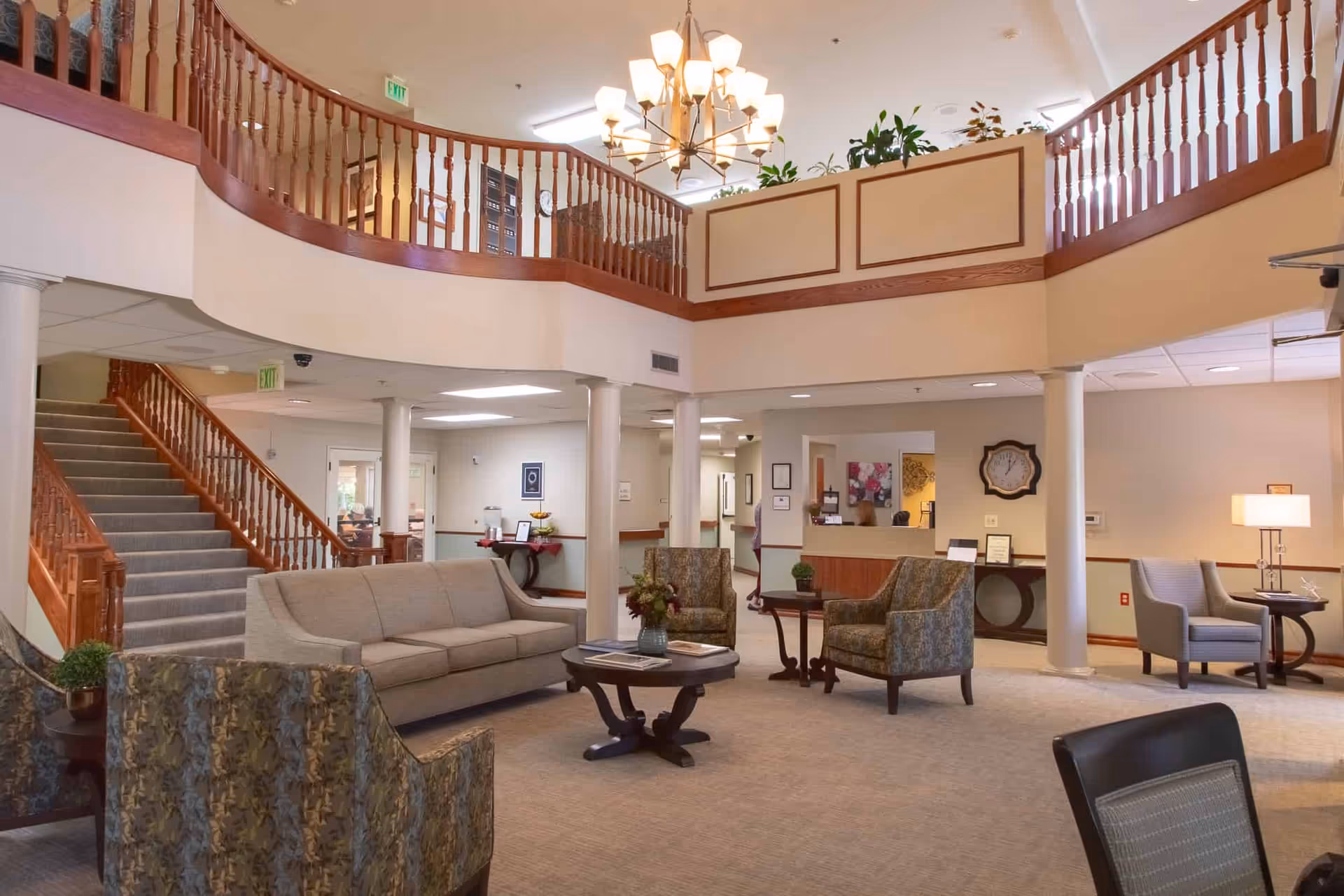 Spacious two-story senior living lobby with seating areas, a staircase and balcony overlooking a reception desk.