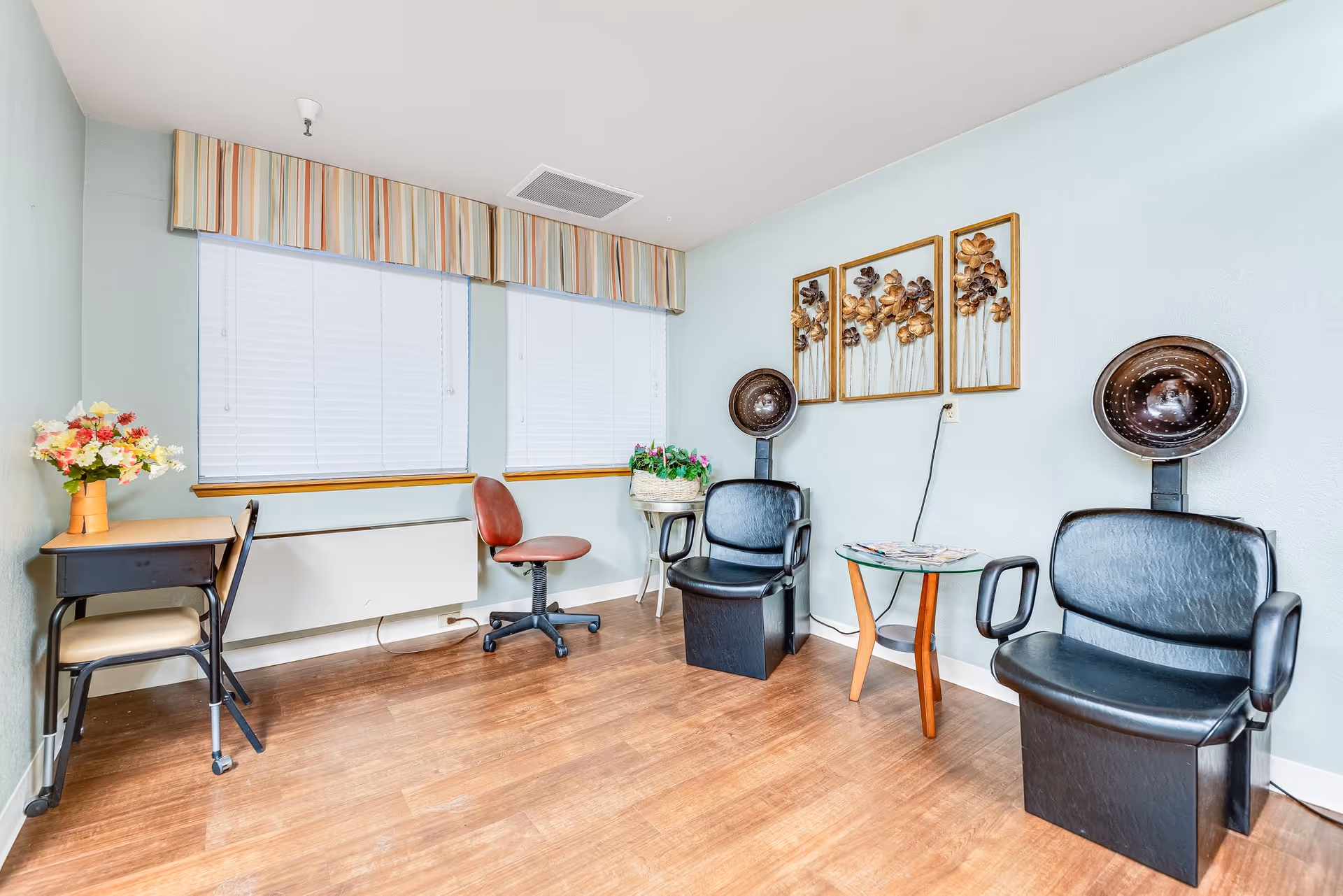A small room with two black salon hair dryer chairs, a red swivel chair, a small glass table with magazines, a small desk with a chair and a vase of flowers, two windows with blinds and striped valances, and wall art featuring metal flowers.