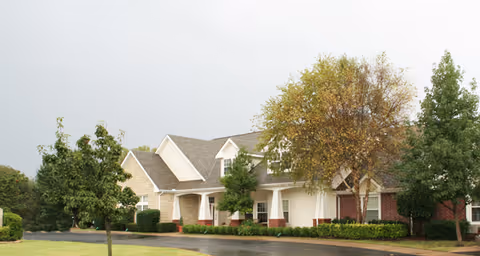 Exterior view of a single-story senior living facility building with a combination of brick and light-colored siding, surrounded by green trees and shrubs under an overcast sky.