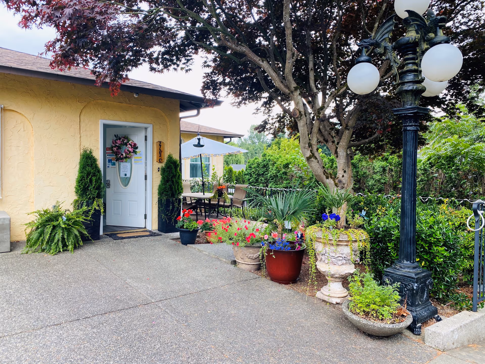 Outdoor view of a facility entrance with a white door decorated with a floral wreath. The building has a yellow textured exterior wall. There are various potted plants and flowers arranged along the walkway, a large tree providing shade, and a black vintage-style street lamp with three round white lights. In the background, there is a patio area with a table, chairs, and an umbrella.