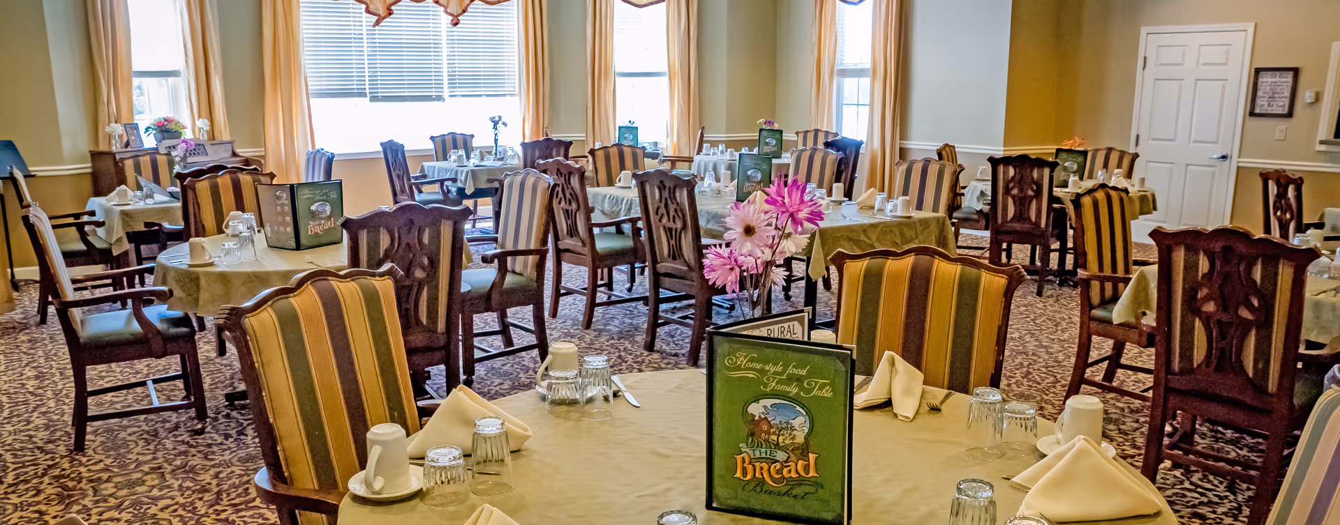 A dining room with multiple round tables covered with beige tablecloths. Each table is set with folded napkins, upside-down glasses, coffee cups, and menus titled 'The Bread Basket'. The chairs have wooden frames with striped upholstery. There are large windows with yellow curtains letting in natural light, and a vase with pink and white flowers is on one of the tables.
