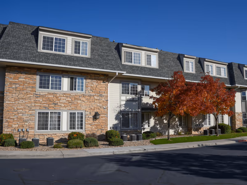 Exterior view of a multi-story senior living facility building with stone and siding facade, multiple windows, and a tree with red autumn leaves in front under a clear blue sky.