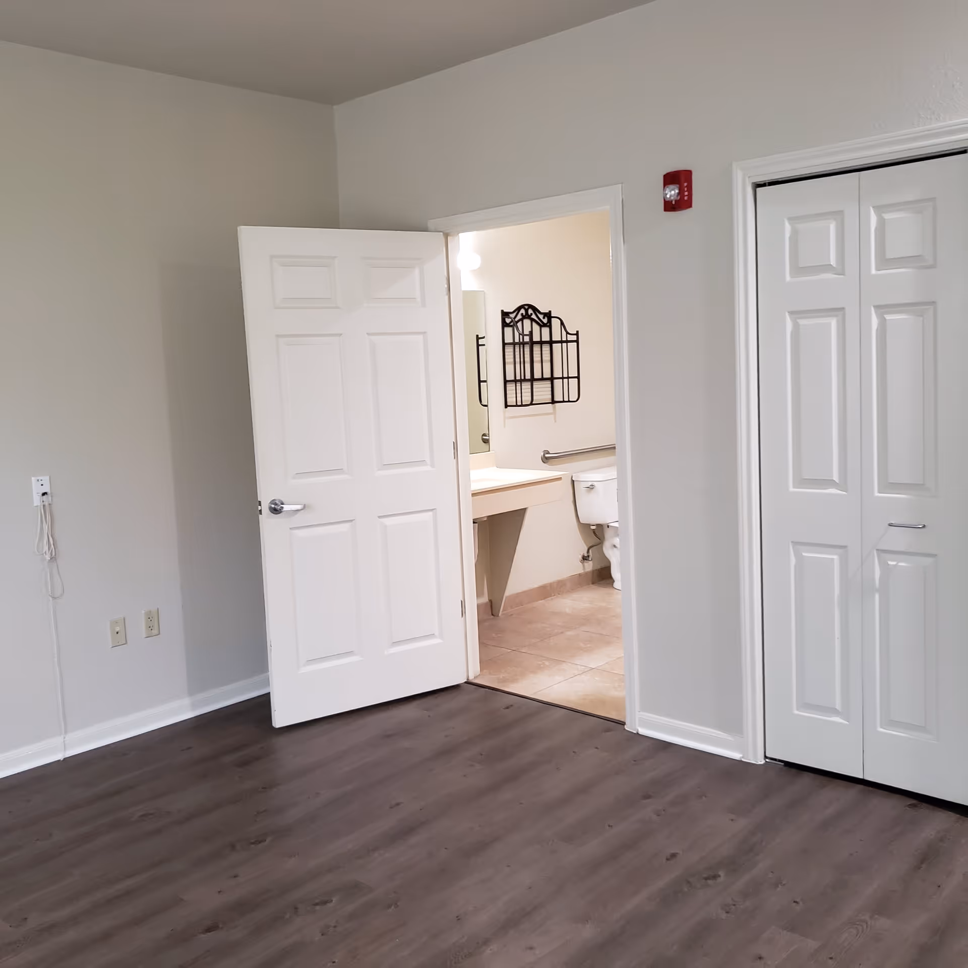 Empty room with wood flooring, white walls, a partially open door revealing a bathroom with a toilet, sink, and mirror, and a closed closet with white double doors.