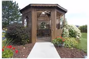 A wooden gazebo with a ceiling fan located in a garden area surrounded by various shrubs and flowers, with a concrete pathway leading up to it.
