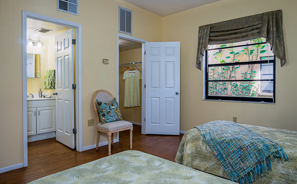 A bedroom with two beds covered in green patterned bedspreads and a blue throw blanket on one bed. There is a wooden chair with a decorative pillow between two open doors. One door leads to a bathroom with a sink, mirror, and towel, and the other door leads to a closet with a hanging dress. A window with a valance shows green plants outside.