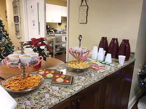 A granite countertop with snacks and beverages arranged for a holiday gathering. There are plates with crackers and cookies, candy canes, holiday-themed napkins, disposable cups, and several burgundy-colored beverage dispensers. A poinsettia plant and a decorated Christmas tree are visible in the background, along with a kitchen area.