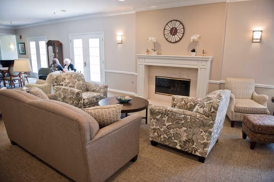 A cozy living room area in a senior living facility with a beige sofa, two floral armchairs, a patterned armchair, and a matching ottoman arranged around a round wooden coffee table. A white fireplace with decorative items and a large clock above it is on the wall. Two elderly women are seated at a table near glass doors in the background.