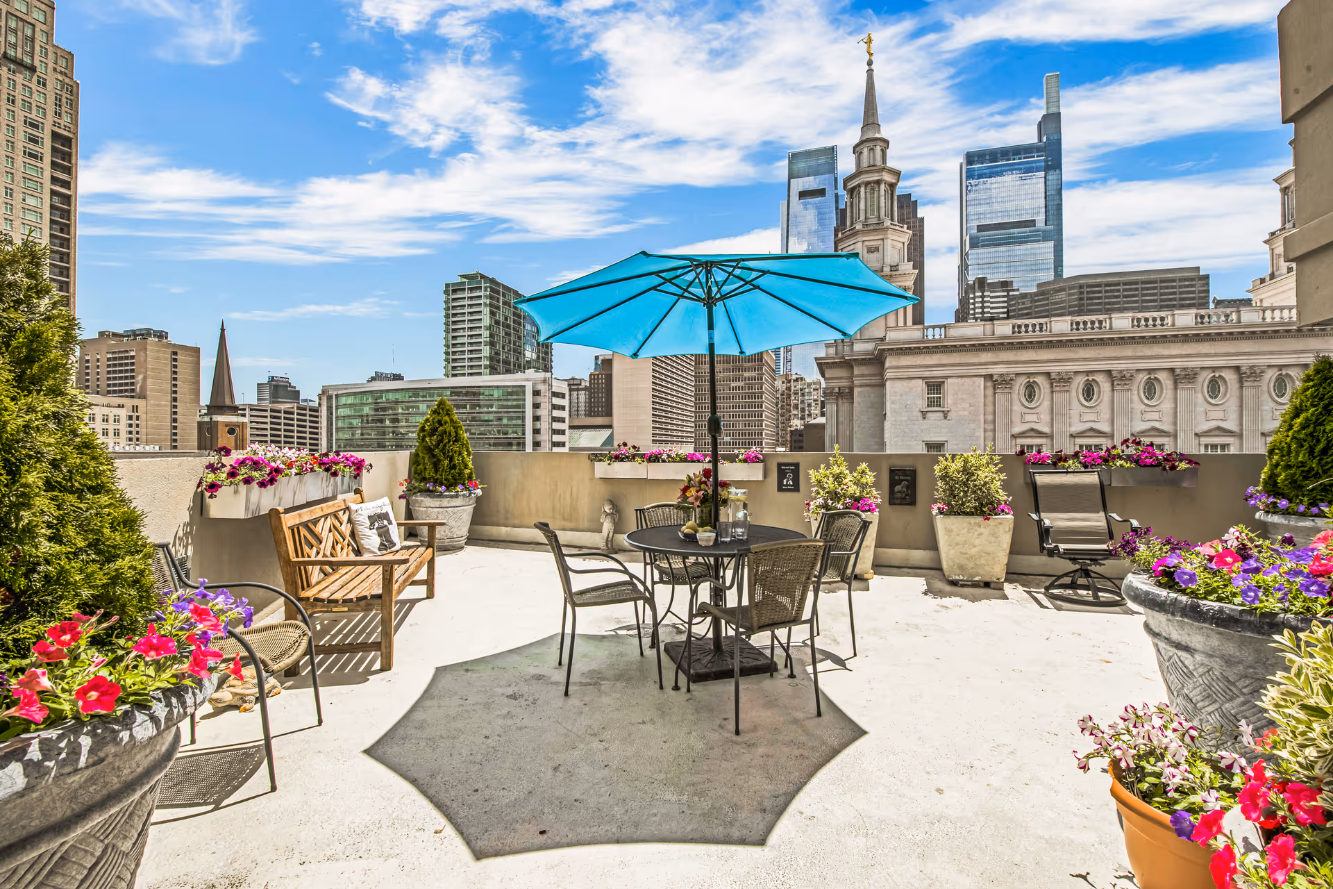 Rooftop terrace with a table, blue umbrella, chairs and potted flowers overlooking a city skyline.