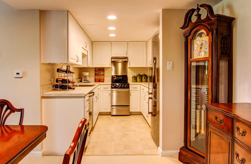 A well-lit kitchen with white cabinets, a stainless steel stove and refrigerator, a countertop with a wine rack and coffee maker, and a wooden dining table with chairs. To the right, there is a tall wooden grandfather clock and a matching wooden sideboard.