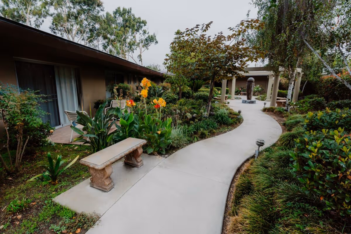 A winding concrete pathway through a well-maintained garden area with various plants, flowers, and trees. There is a stone bench on the left side of the path and a covered seating area with columns and a sculpture in the background. The garden is adjacent to a building with sliding glass doors and patio chairs.