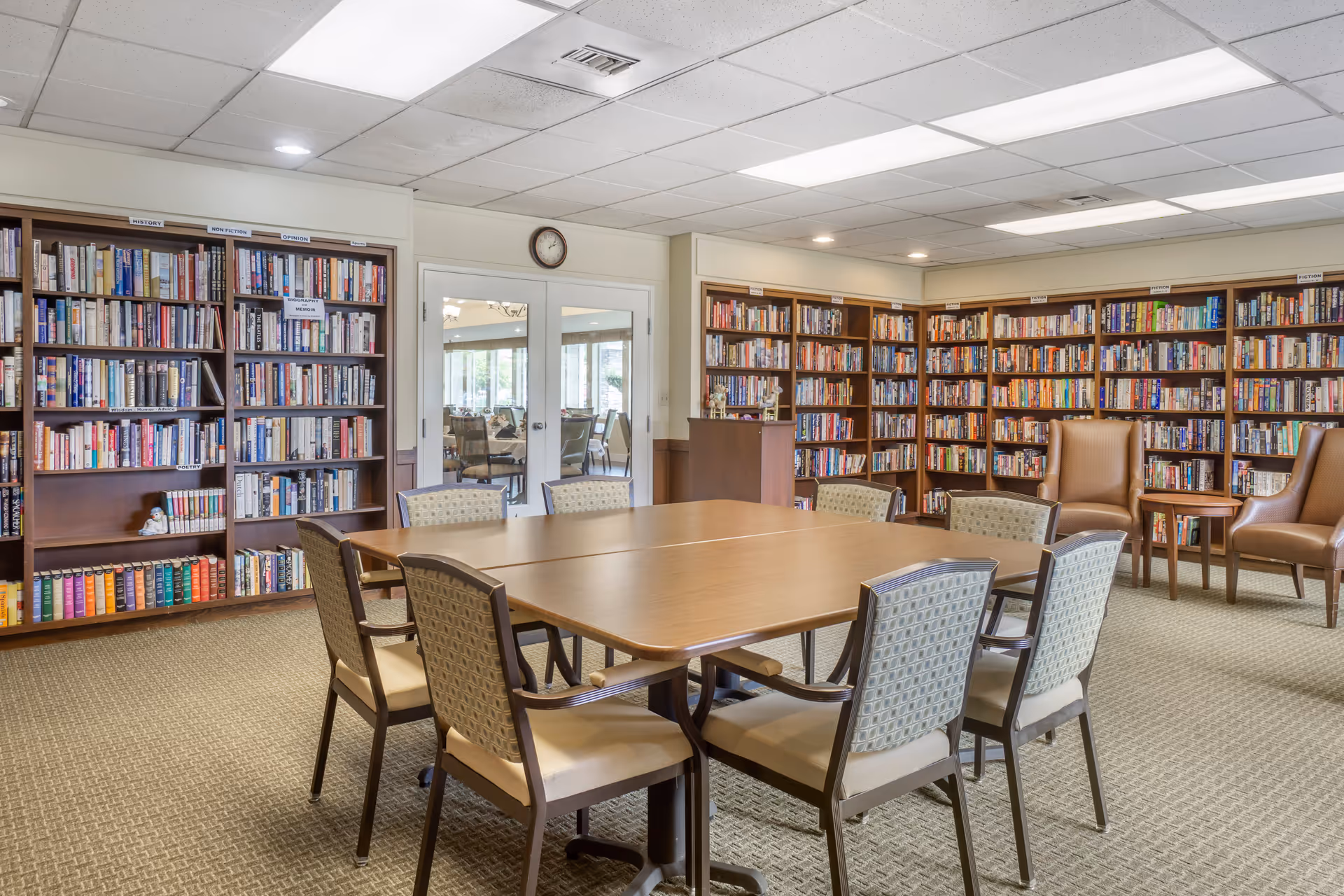 A well-lit library room with bookshelves filled with books lining the walls. In the center, there are two wooden tables pushed together surrounded by eight cushioned chairs. Two brown leather armchairs and a small round table are positioned near the bookshelves. A clock is mounted above double glass doors leading to another room.