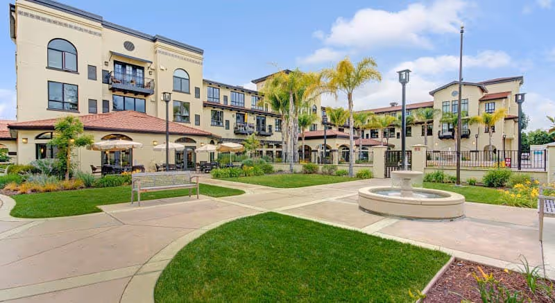 Outdoor courtyard area of a senior living facility with a central water fountain, green lawns, palm trees, benches, and a three-story building with balconies and large windows in the background under a partly cloudy sky.