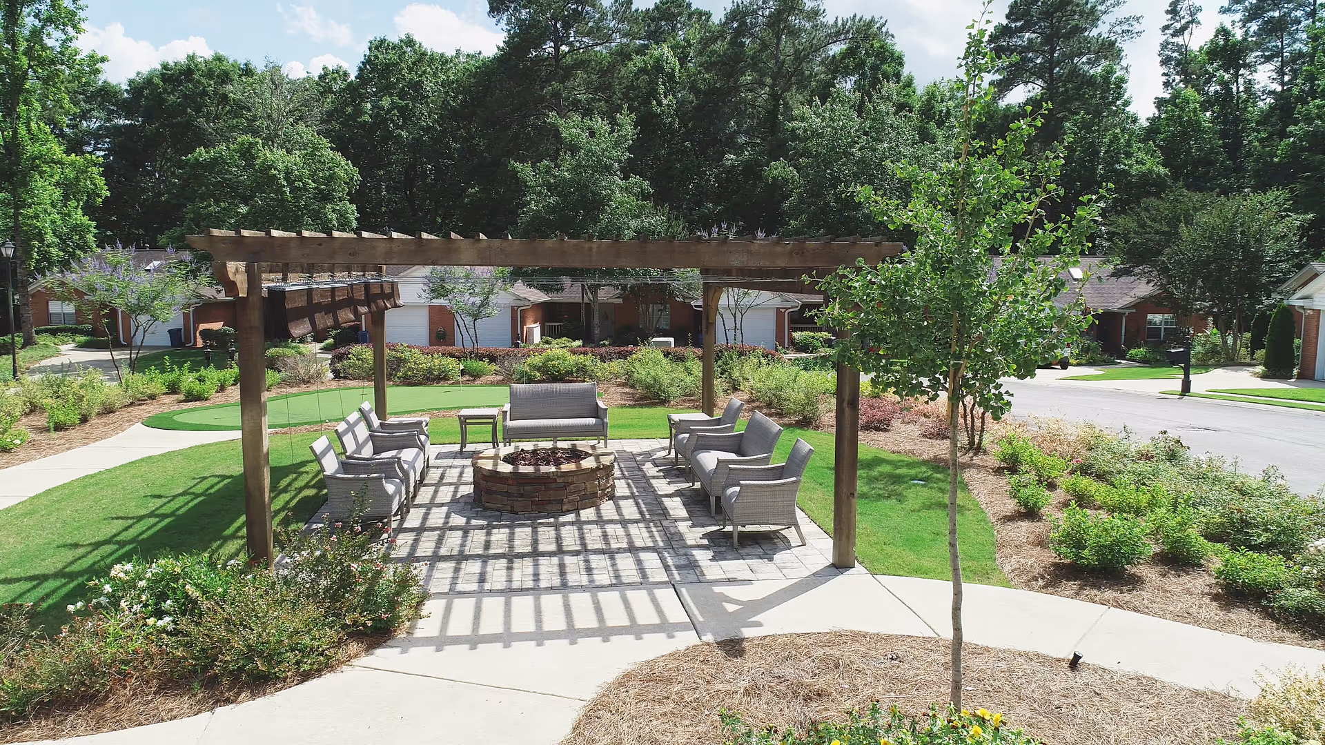 Outdoor seating area with a wooden pergola casting shadows on the ground, surrounded by cushioned chairs and a stone fire pit in the center. The area is landscaped with green grass, bushes, and trees, with residential buildings visible in the background under a partly cloudy sky.