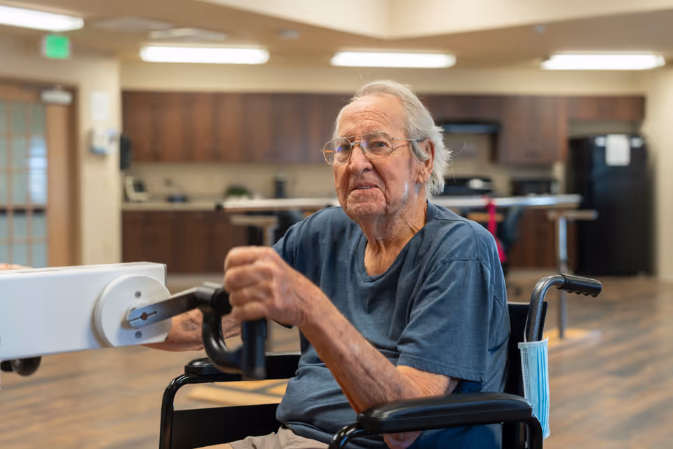 An elderly man in a wheelchair using a hand-crank exercise device in a communal room with a kitchenette in the background.