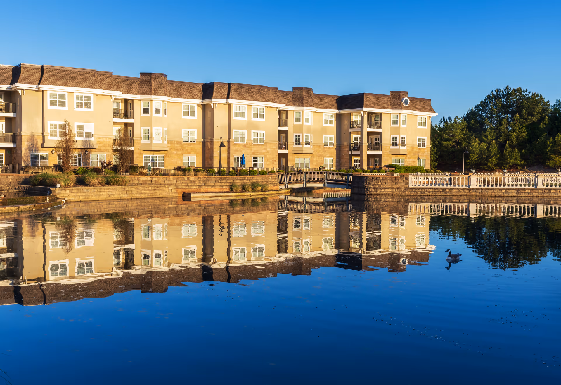 A large, multi-story beige and brown retirement community building reflected in a calm body of water with a clear blue sky above. There is a small bridge and a duck swimming in the water near the building. Trees are visible in the background.