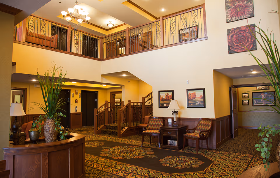 Interior view of a senior living facility lobby with a two-story ceiling, decorative railing on the upper level, patterned carpet, wooden furniture including chairs and tables, potted plants, framed artwork on the walls, and warm lighting fixtures.