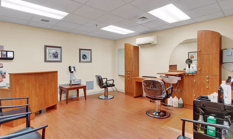 Interior view of a salon area in a senior living facility with wooden flooring, two salon chairs, a wash basin, wooden cabinets, a small table with a telephone, and framed pictures on the wall under fluorescent ceiling lights.
