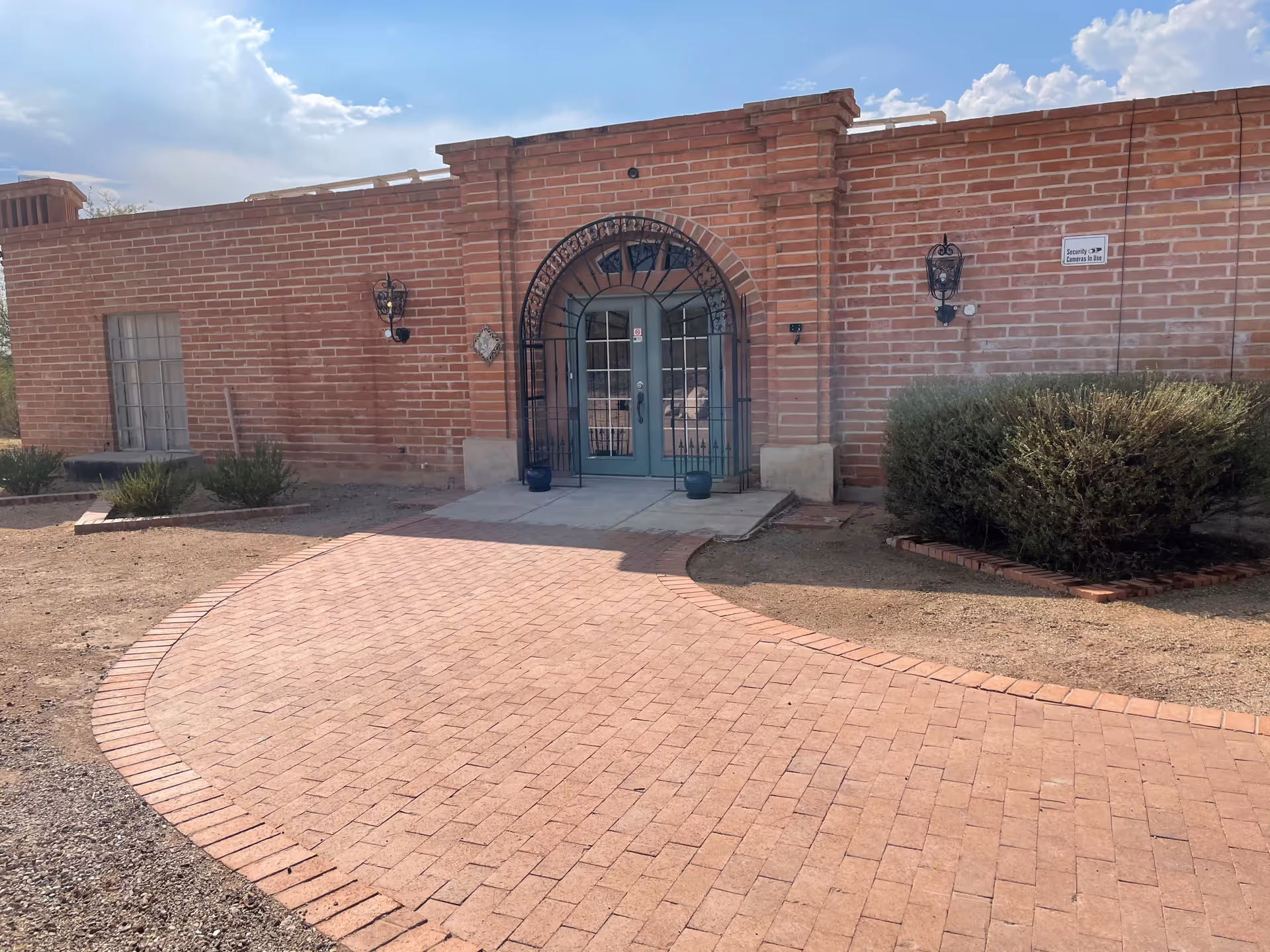 Front entrance of a brick building with a curved brick pathway leading to a double door with an arched wrought iron gate. There are two wall-mounted lantern-style lights on either side of the door and some bushes and plants near the building. A sign on the wall reads 'Security Cameras In Use'.