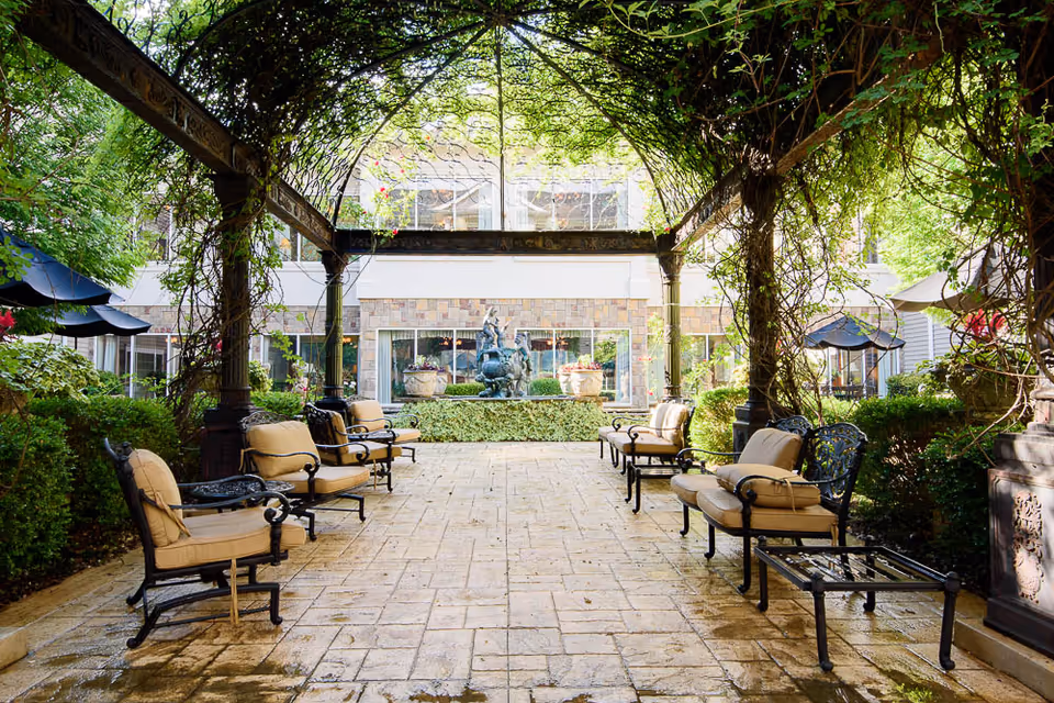 Outdoor seating area with cushioned metal chairs arranged under a pergola covered with greenery. In the background, there is a building with large windows and a decorative fountain surrounded by plants.