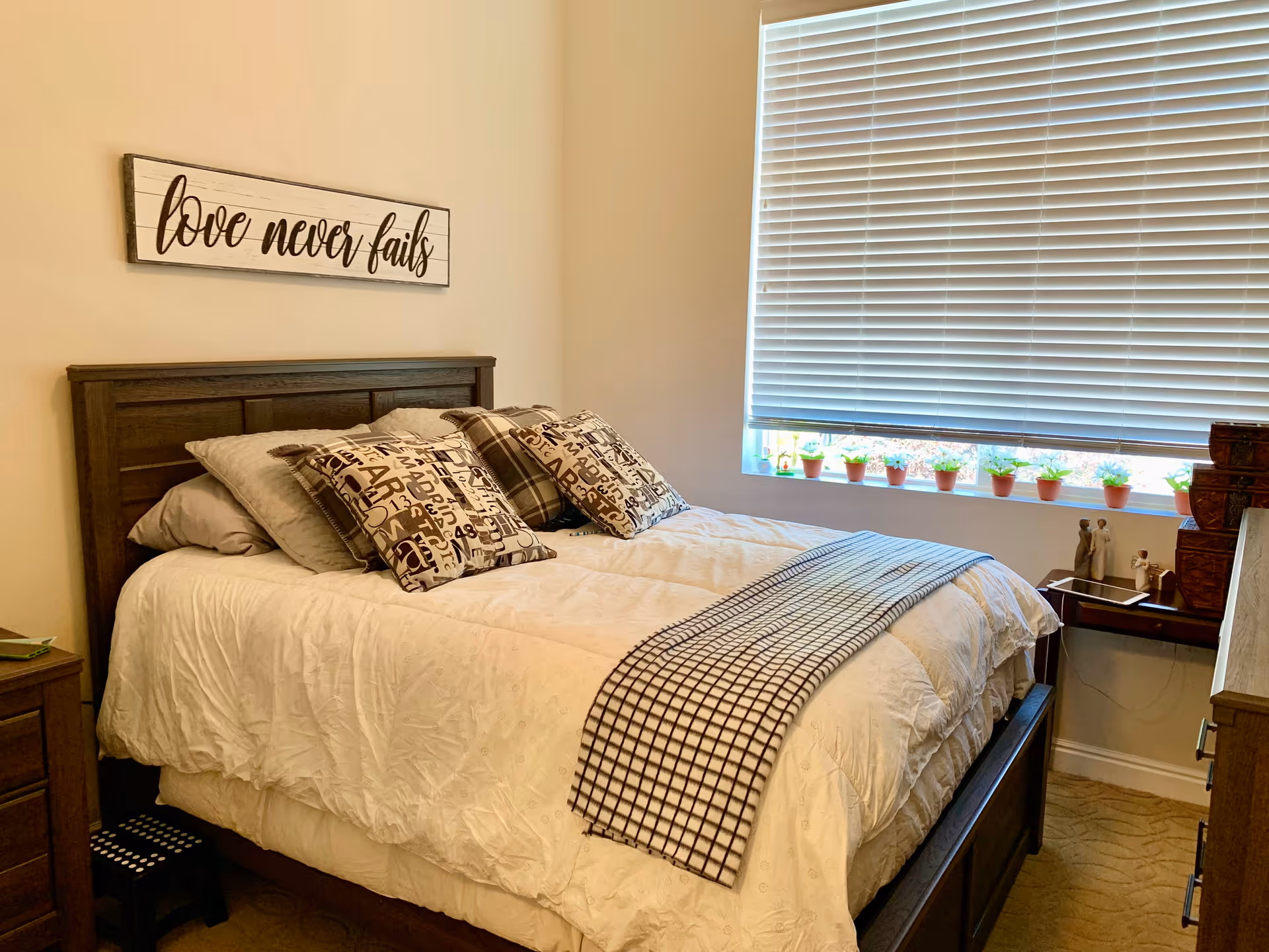 A cozy bedroom with a wooden bed frame, white bedding, and several decorative pillows. Above the bed is a wall sign that reads 'love never fails'. A window with closed blinds is on the right side, with small potted plants on the windowsill. There is a nightstand with a few decorative items and a small step stool beside the bed.