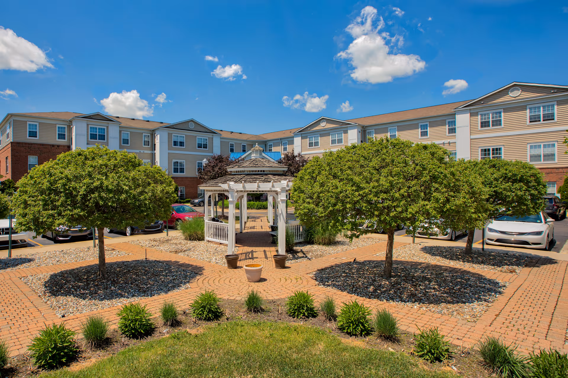 Outdoor courtyard with a white gazebo, paved walkways, trimmed trees, and a multi-story residential building behind it under a blue sky.