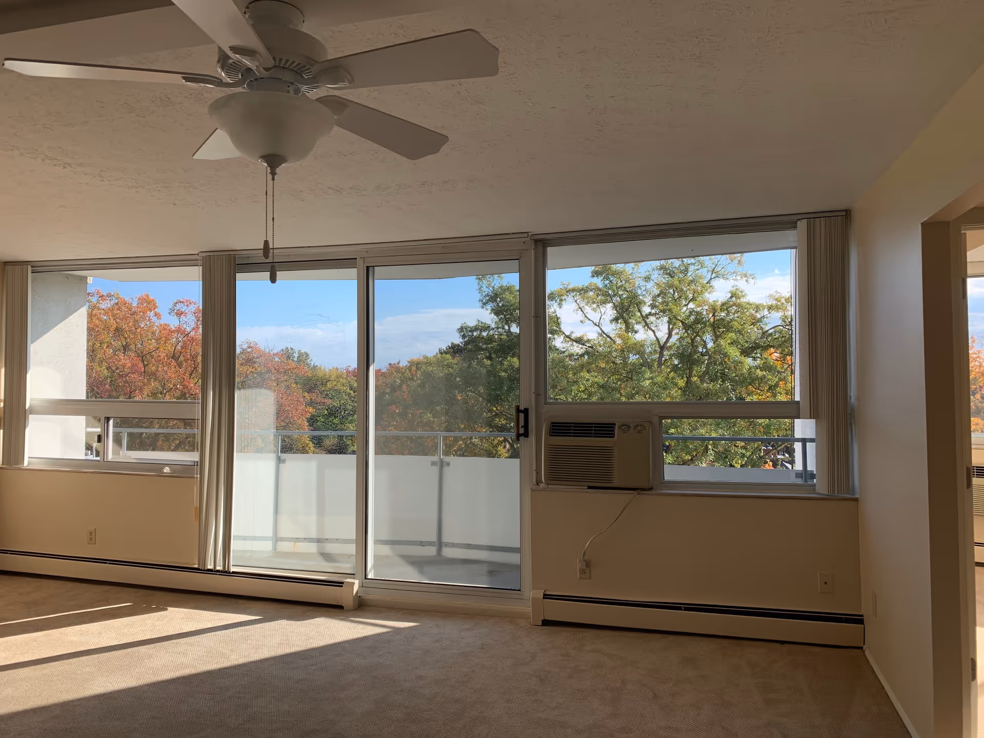 An empty living room with beige carpet and cream-colored walls featuring large windows and a sliding glass door leading to a balcony. The windows show a view of trees with autumn foliage. A white ceiling fan with light is mounted on the ceiling, and a window air conditioning unit is installed in one of the windows.