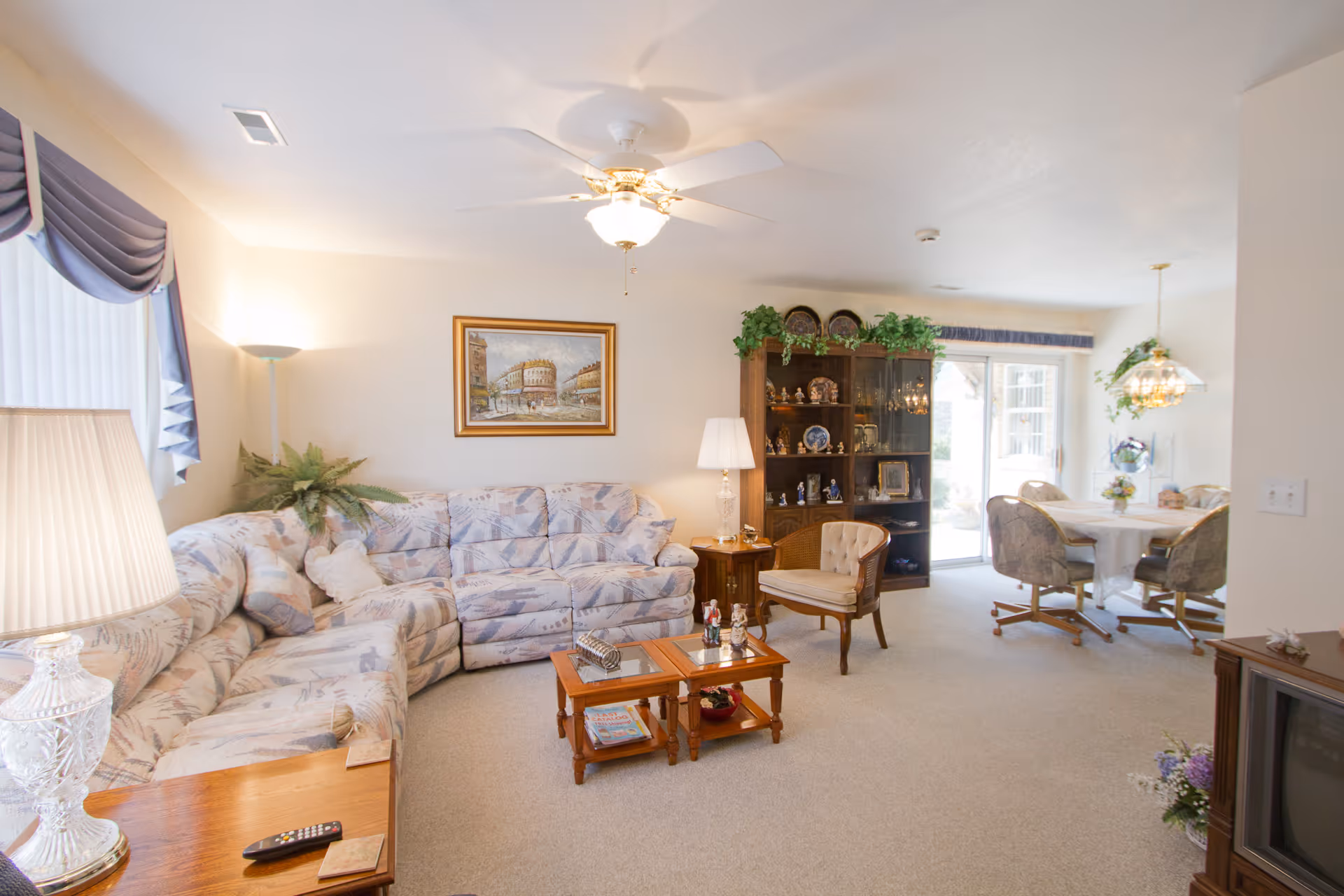 Bright carpeted living area with a patterned sectional sofa, coffee tables, a display cabinet, and an adjacent dining nook.