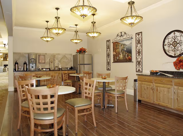 A dining area with several round tables and wooden chairs with green cushions. The room features hanging decorative light fixtures, a wooden floor, and a kitchenette area with a refrigerator, coffee machine, and cabinets. Wall decorations include framed artwork and ornamental metal pieces.