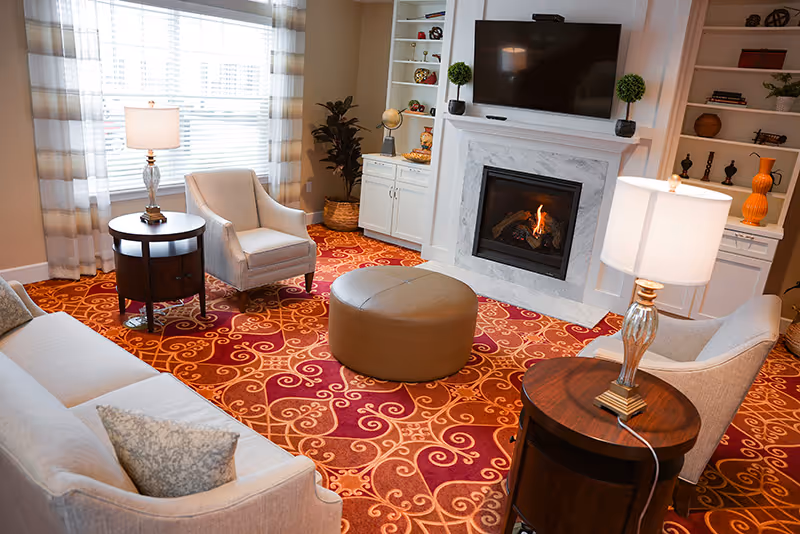 A cozy living room with a patterned red and orange carpet, a beige sofa with a decorative pillow, two beige armchairs, two round wooden side tables with lamps, a round brown ottoman in the center, a white fireplace with a fire burning, a mounted flat-screen TV above the fireplace, and built-in white shelves with decorative items.