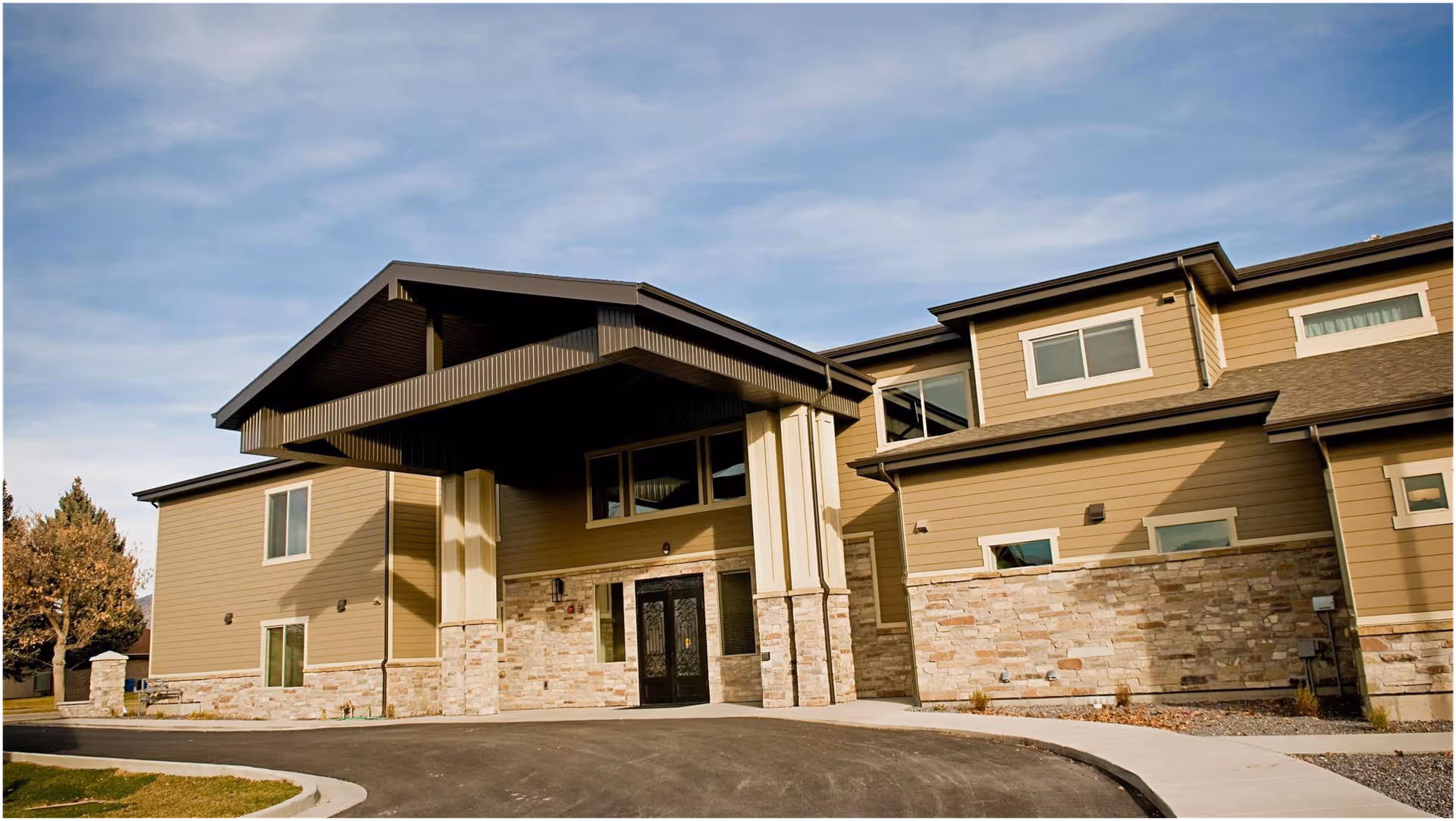 Exterior view of a modern assisted living facility building with beige siding and stone accents, featuring a covered entrance with large pillars and a driveway in front under a partly cloudy sky.