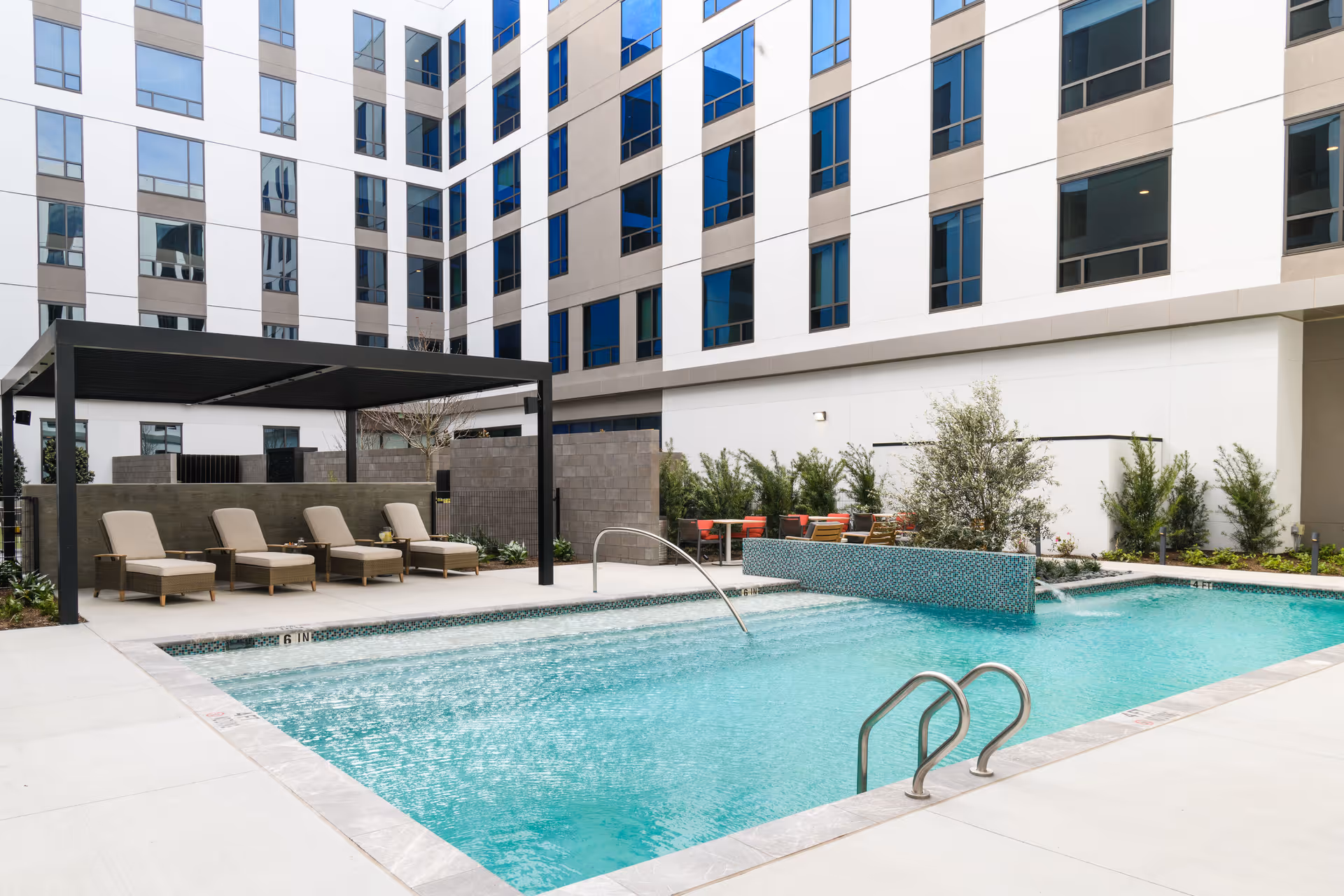 Outdoor swimming pool area at a senior living facility with lounge chairs under a shaded pergola, a water feature in the pool, and a modern multi-story building in the background.