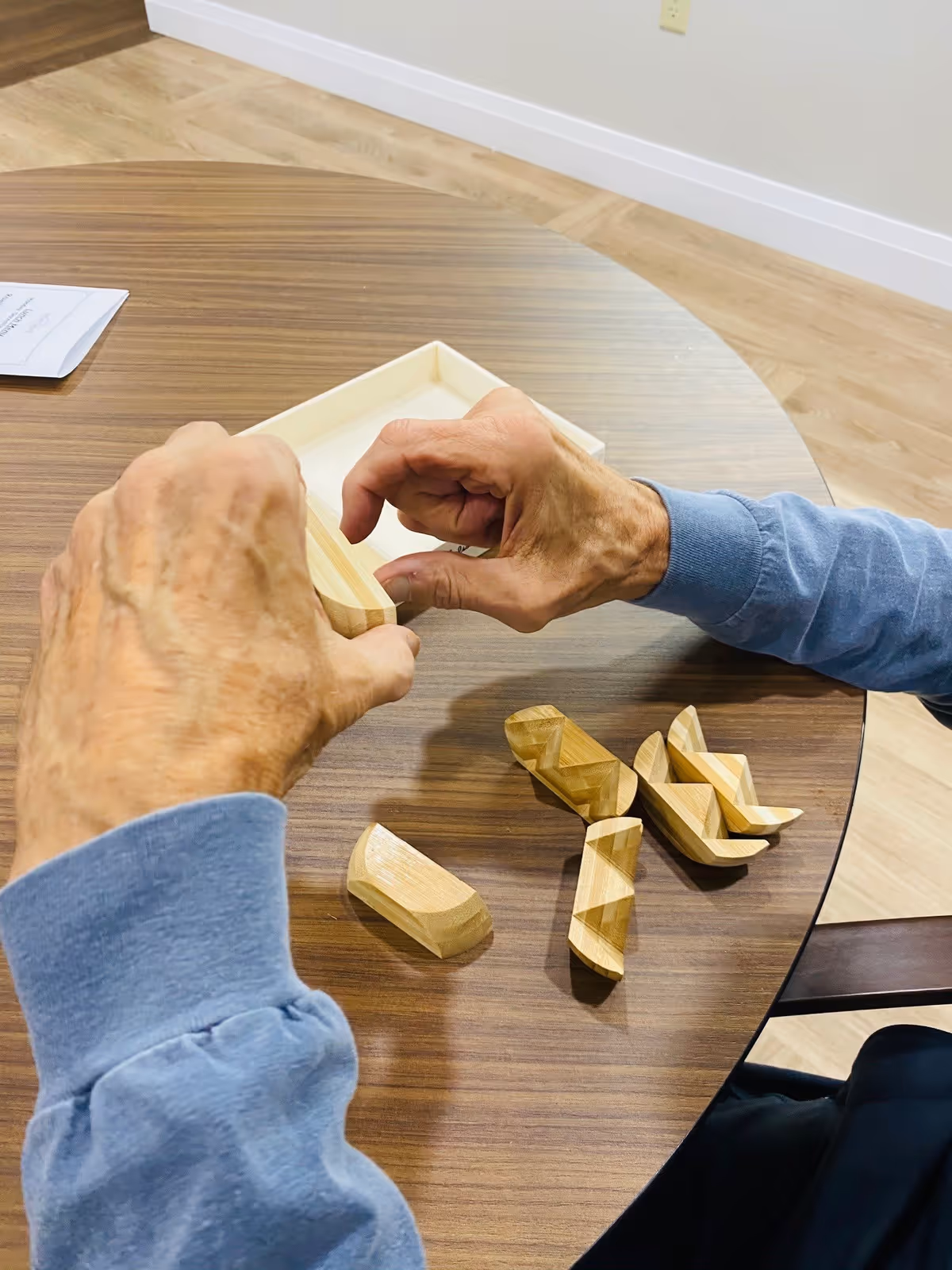 An elderly person wearing a blue long-sleeve shirt is assembling wooden puzzle pieces on a round wooden table with a light wood floor in the background.