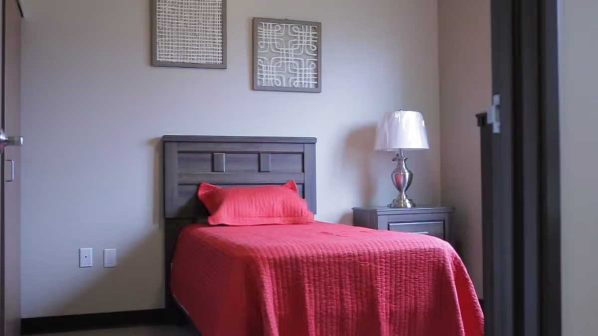 A bedroom with a single bed covered in a red quilt and matching pillow. The bed has a dark wooden headboard. Next to the bed is a dark wooden nightstand with a silver lamp on it. Two framed abstract artworks hang on the beige wall above the bed.