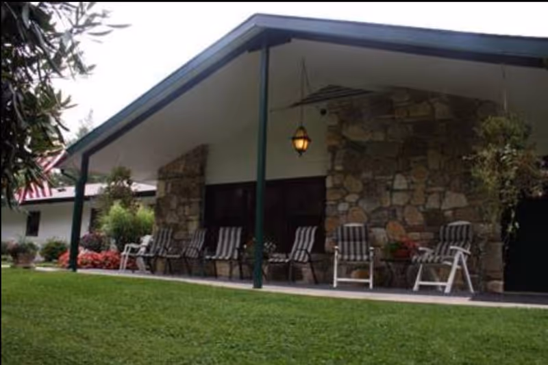 Outdoor patio area of a building with a stone wall and a slanted roof. Several striped cushioned chairs are arranged along the patio, with hanging plants and a lantern light fixture. The foreground shows a well-maintained green lawn.