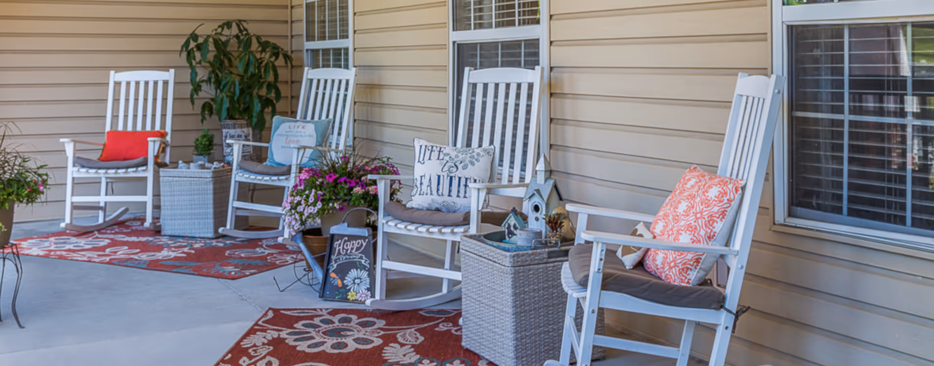 A covered outdoor patio area with four white rocking chairs, each with cushions and decorative pillows. The patio floor has red patterned rugs, and there are potted plants and small decorative items placed around the seating area. The background shows beige siding and windows with white blinds.