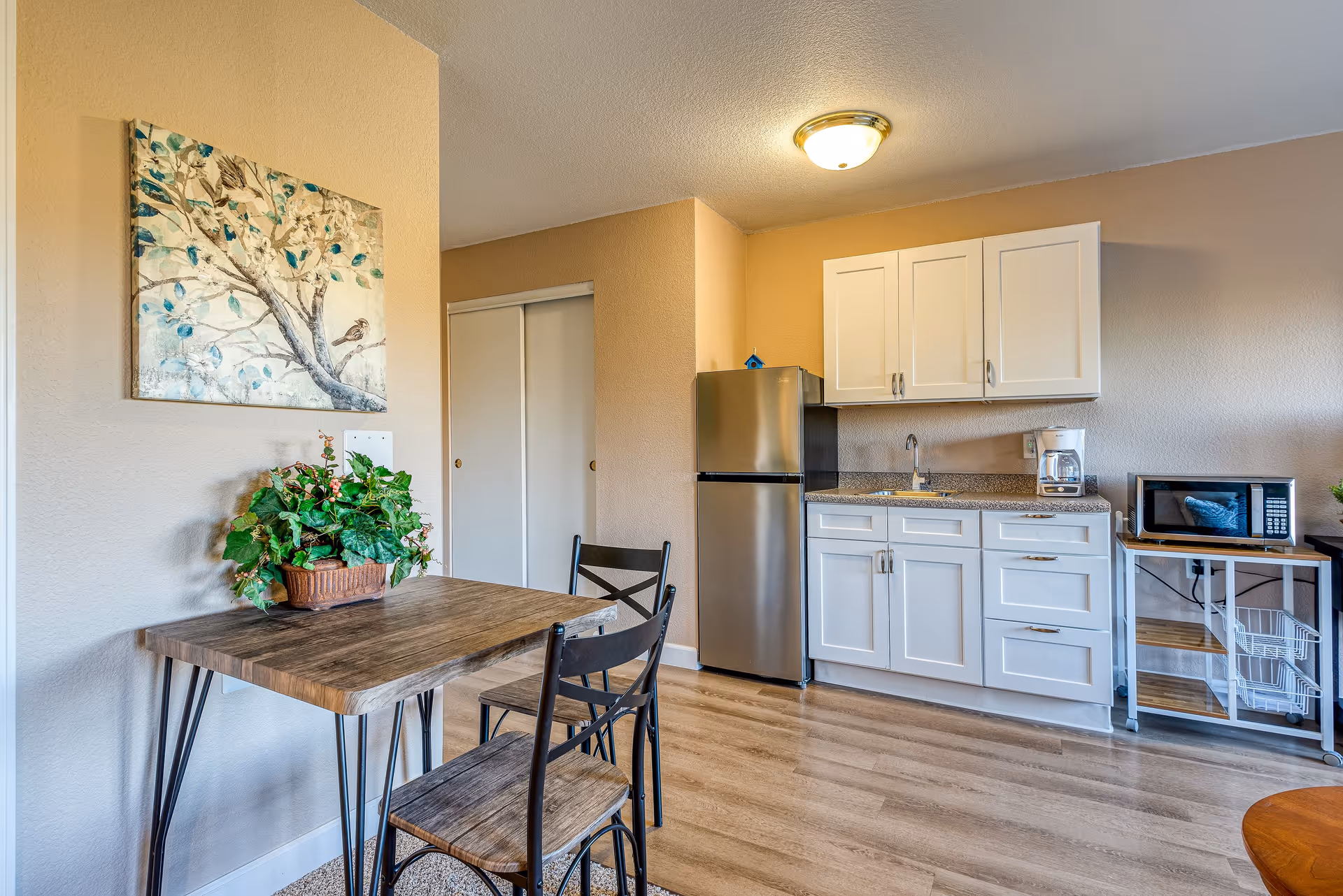 A small kitchen area with white cabinets, a stainless steel refrigerator, a microwave on a metal cart, and a coffee maker on the countertop. In front of the kitchen is a wooden table with two chairs and a bench, decorated with a potted green plant. A painting of a tree with birds hangs on the beige wall above the table.