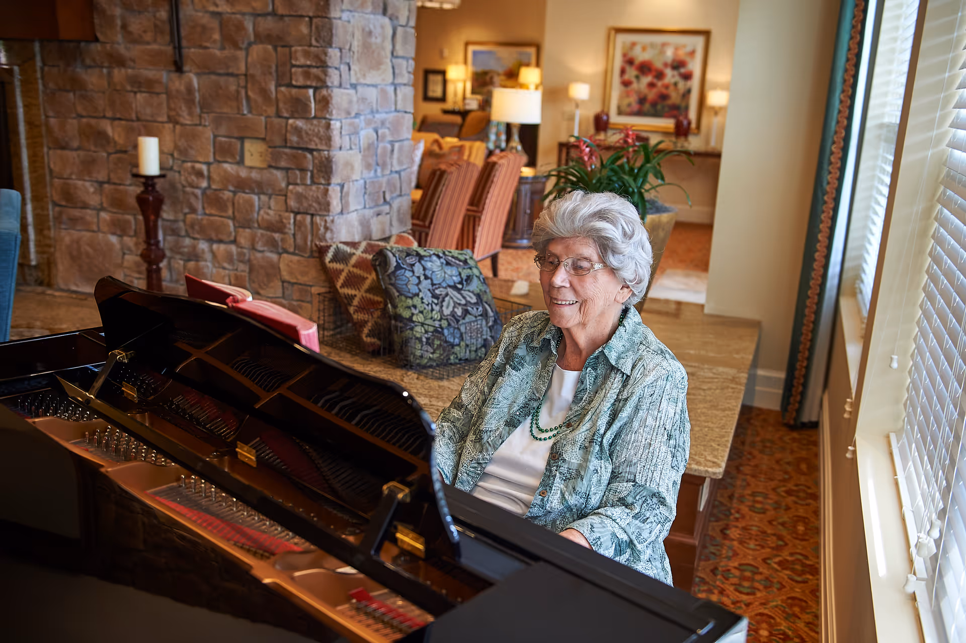 An elderly woman with gray hair and glasses is sitting at a grand piano in a cozy, well-lit living area. The room features a stone wall, patterned cushions, a plant on a countertop, and a window with blinds letting in natural light. In the background, there are chairs, a lamp, and framed artwork on the walls.