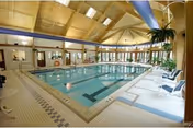 Indoor swimming pool area with clear water, surrounded by tiled flooring and several lounge chairs. The ceiling is high with wooden beams and skylights allowing natural light to brighten the space. Large windows line the far wall, providing a view of greenery outside.