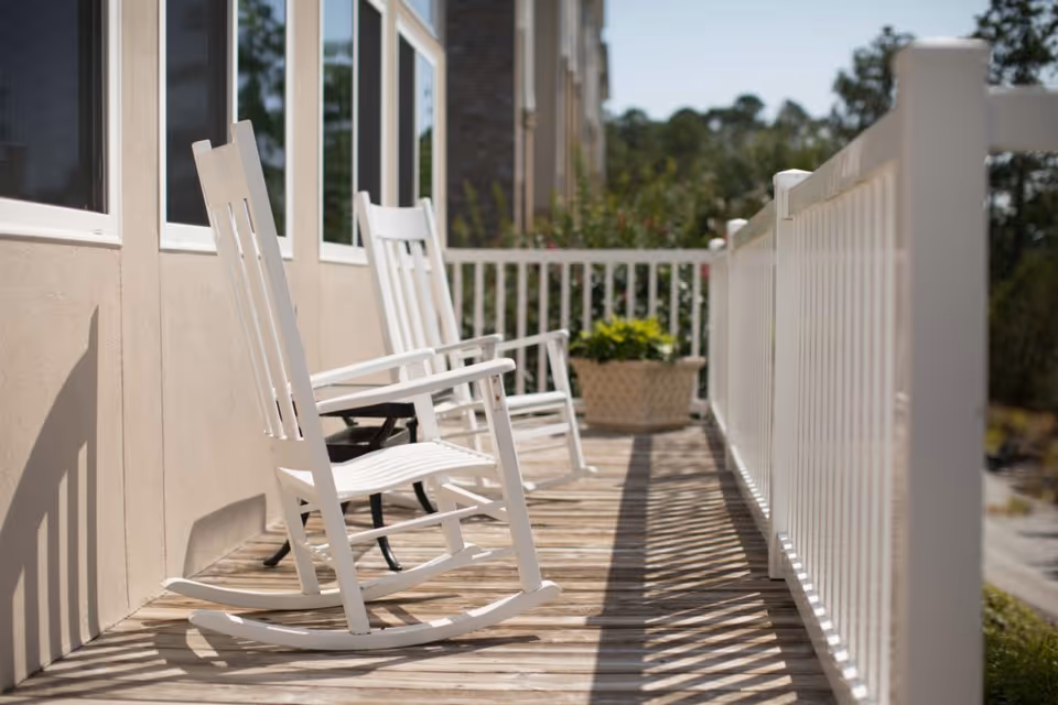 A wooden porch with white railings featuring two white rocking chairs and a planter with green plants at the far end, adjacent to a building with several windows.