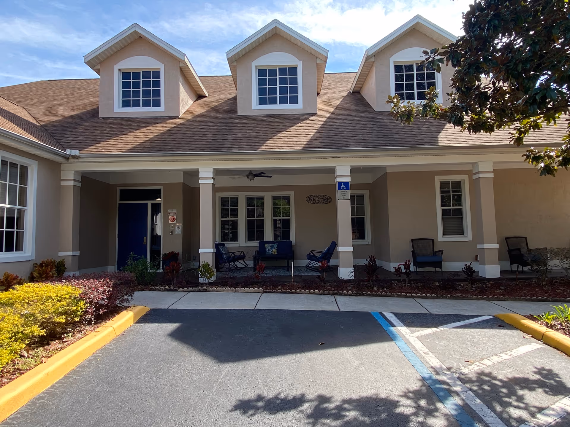Front exterior view of a beige assisted living facility with three dormer windows on the roof, a covered porch with seating, and a blue entrance door. There is a handicapped parking space in front and landscaped bushes along the walkway.