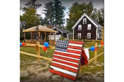 Outdoor area with a wooden fence decorated with red, white, and blue balloons. In front of the fence is a wooden board painted with the American flag design. Behind the fence, there is a gazebo and a two-story dark brown house with white trim surrounded by trees.