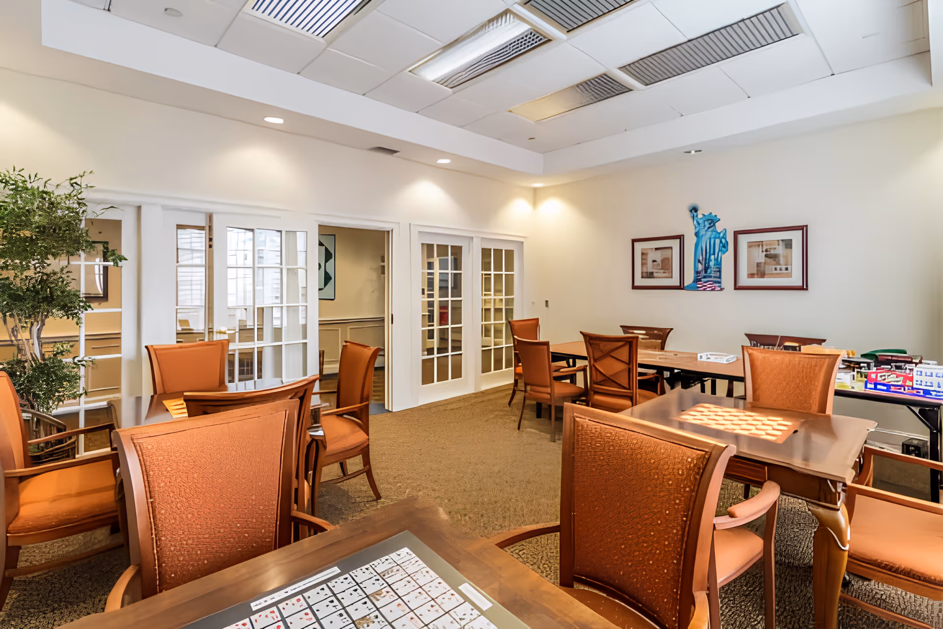 A well-lit room with several wooden tables and orange cushioned chairs arranged for group activities. On the tables, there are board games and card games. The room has beige walls, carpeted floor, and white ceiling tiles with recessed lighting. There are framed pictures on the walls and glass-paneled double doors leading to another room.