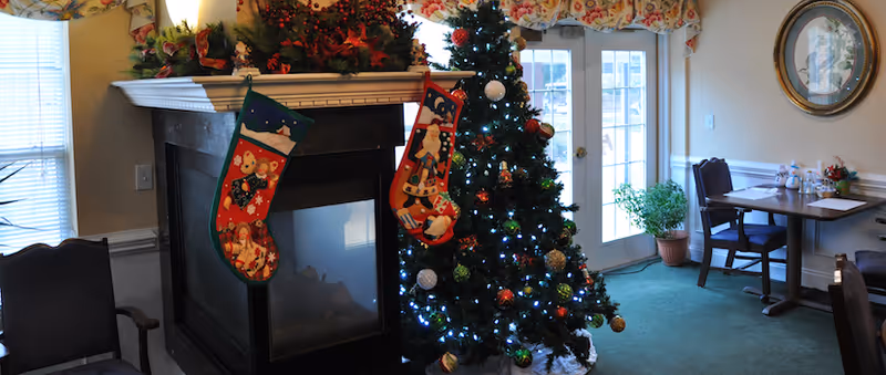 A cozy room decorated for Christmas with a decorated Christmas tree adorned with lights and ornaments next to a fireplace with two Christmas stockings hanging from the mantel. There are chairs and a small table near a glass door with a potted plant beside it. The room has green carpet and floral curtains above the door.