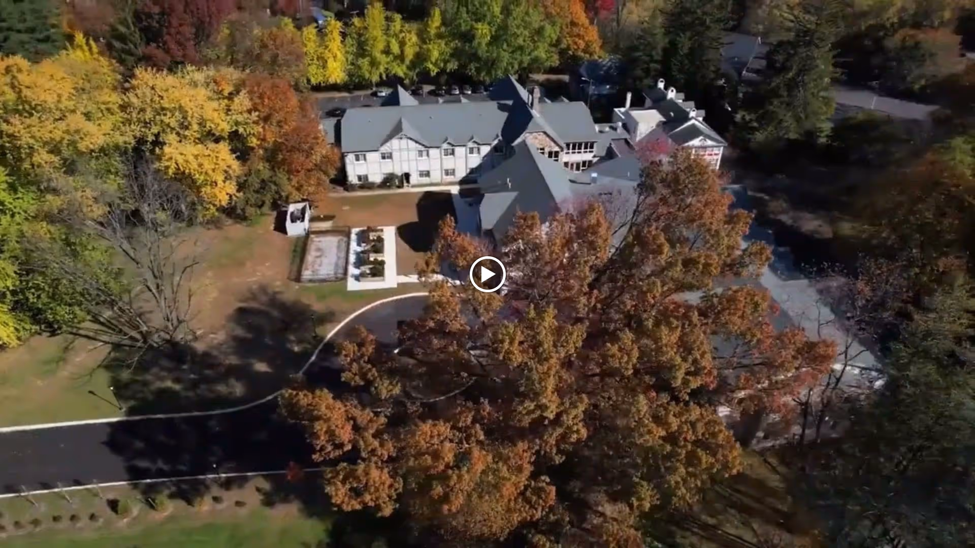 Aerial view of Ivy Gables Senior Living facility surrounded by trees with autumn foliage. The building has multiple sections with gray roofs and white walls. There is a curved driveway leading to the entrance and a garden area visible on the left side.