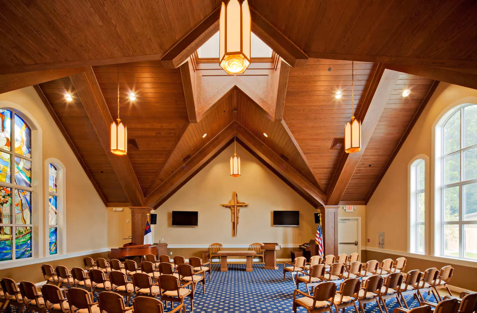 Interior view of a chapel with a wooden vaulted ceiling, rows of chairs, stained glass windows, an altar with a crucifix, and pendant lights.