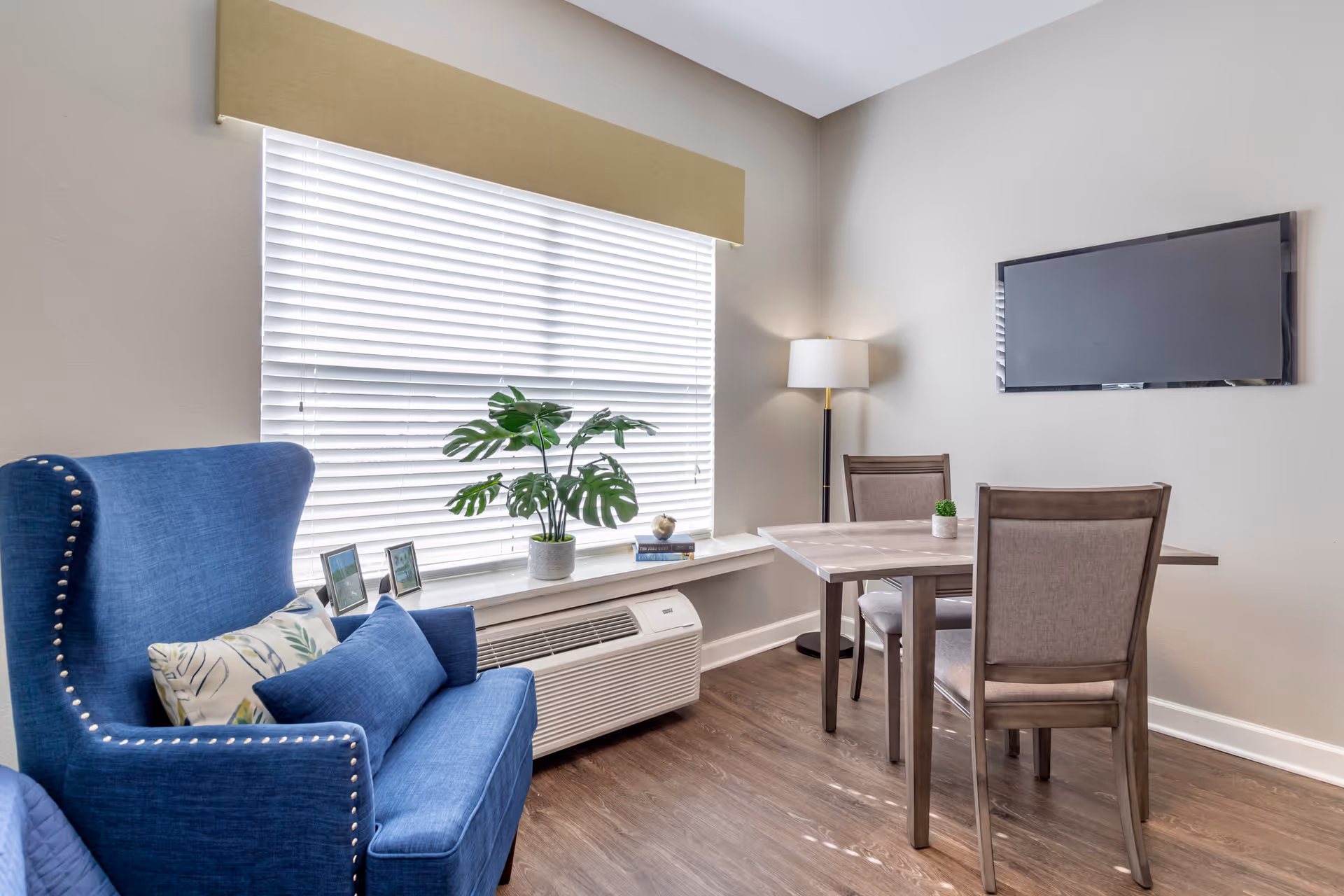 Bright sitting area with a blue wingback chair, a small dining table, and a wall-mounted TV beside a large window.