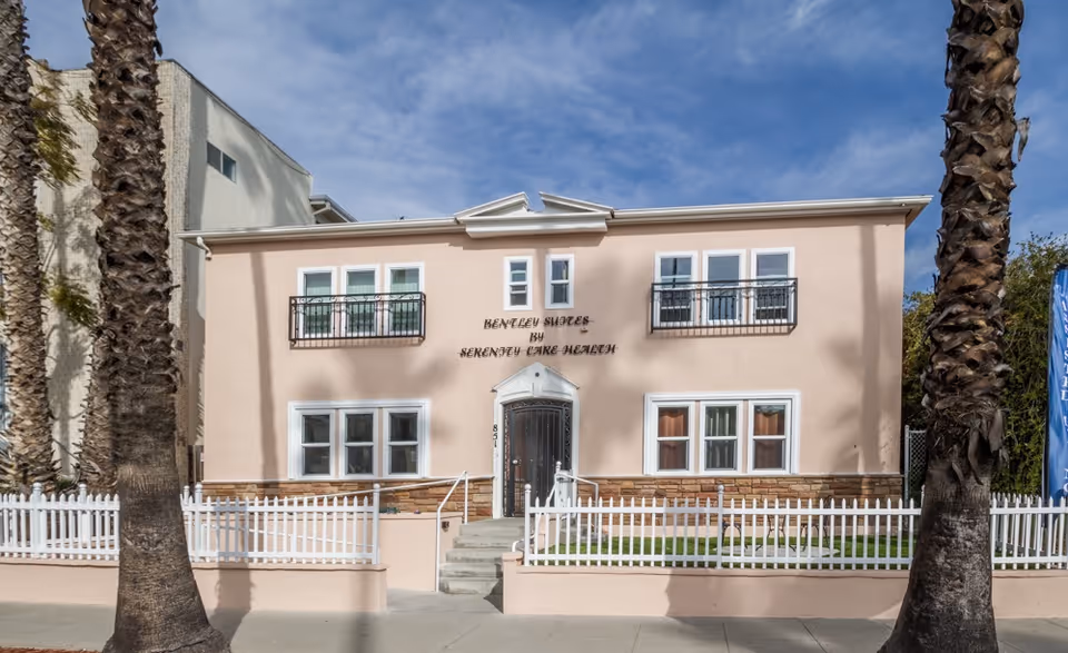 Front exterior view of a two-story building with a light pink facade and white trim, labeled Bentley Suites by Serenity Care Health. The building has several windows with black railings on the upper floor and a white picket fence surrounding a small lawn area. Two tall palm trees are visible in front of the building under a partly cloudy sky.